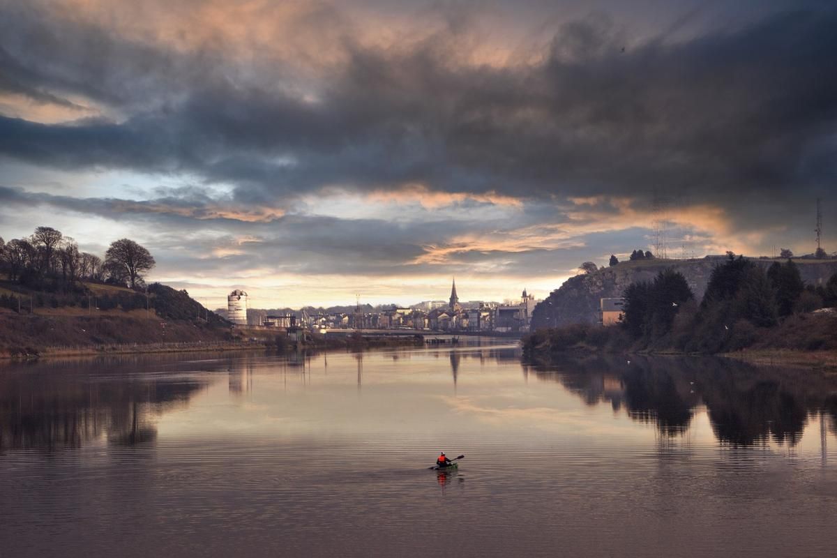 Kayakiste pagayant seul sur une rivière calme avec une ville et ses églises en arrière-plan au coucher du soleil Kayakiste pagayant seul sur une rivière calme avec une ville et ses églises en arrière-plan au coucher du soleil