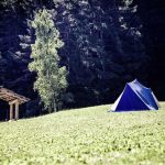 Tente bleue sur une colline herbeuse, abri en bois à gauche, forêt dense en arrière-plan
