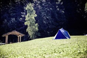 Tente bleue sur une colline herbeuse, abri en bois à gauche, forêt dense en arrière-plan