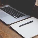 Un ordinateur, un cahier vierge et un stylo reposent sur un bureau en bois